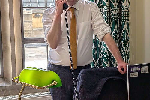Will Forster speaking on the phone in his Westminster office, he is wearing a white shirt with the sleeves rolled up and a yellow tie.