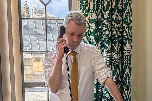 Will Forster speaking on the phone in his Westminster office, he is wearing a white shirt with the sleeves rolled up and a yellow tie.