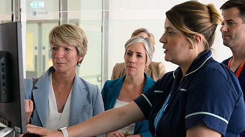 A nurse at Addenbrookes Hospital demonstrates something on a screen to Pippa Heylings, Lib Dem Parliamentary Candidate for South Cambs. Daisy Cooper MP,  the national Liberal Democrat Health and Social Care spokesperson, sits behind Pippa looking at the same screen.