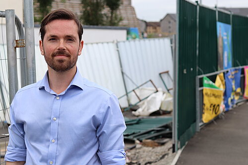 Adam Harley standing in front of an area partly fenced off with rubble and other construction materials in a disorganised fashion.