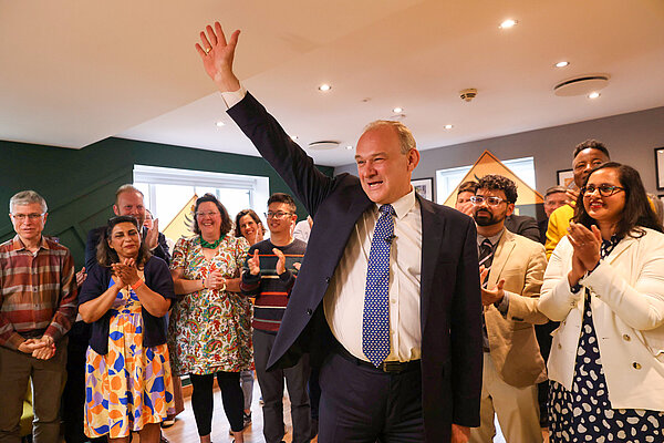 Ed Davey, dressed in a dark suit with blue tie, waves. Behind him party activists applaud Ed.