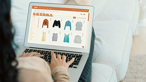 Person sat on a white sofa, back of their shoulder, their hands and their legs visible. The person is using a laptop. On the laptop screen is an online clothes shopping website.