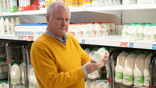 Clive Jones in the milk aisle of a supermarket, looking unimpressed at the price of a pint of milk.