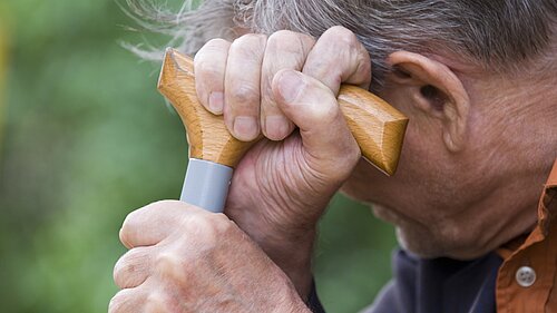 An elderly man holding a walking stick which he is leaning his head on.