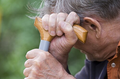 An elderly man holding a walking stick which he is leaning his head on.