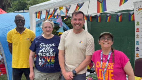 Sean in stood in a group of people (including councillor Liz Needham to his right) in front of a gazebo decorated with Pride flag bunting.  