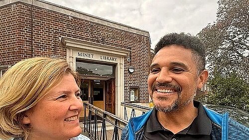 A man and a woman smiling, standing out doors in front of a brick library