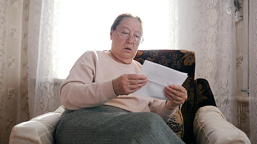 Older woman sat in arm chair looking in shock at a bill.