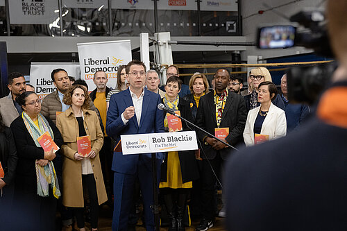 Rob Blackie pictured in a white shirt with a navy blazer. Lib Dem activists pictured behind. 