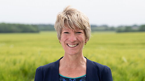 Head and shoulders photo of Pippa Heylings, Lib Dem Parliamentary candidate for South Cambs. Pippa is smiling at the camera, standing in front of a grassy field with trees in the distance.