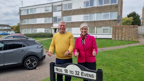 Two people (Tony on the left, Daisy on the right) are standing in front of a block of flats, and behind a sign displaying flat numbers. They are both smiling, looking directly at the camera, and giving a thumbs up.
