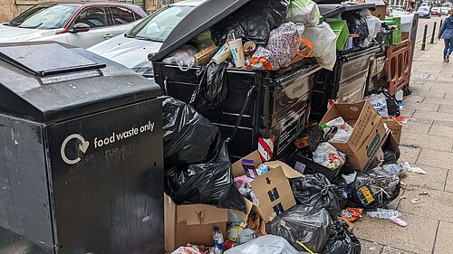 Excess rubbish piled around large bins.