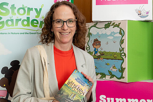 Alison holding the book 'Wildlands' by Brogen Murphy in front of banners promoting the The Reading Agency’s 2025 Summer Reading Challenge.