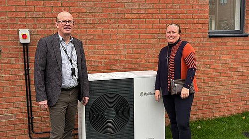 Cllr Paul de Kort and Cllr Jacqui Taylor are standing on either side of the air source heat pumps at the new properties. They are both smiling and looking at the camera, and are stood in front of the brick wall of one of the new properties. 