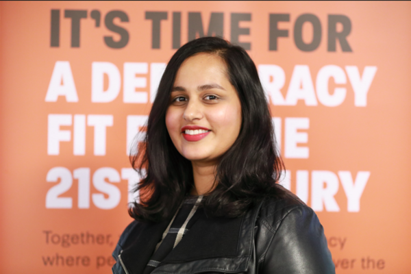 Lib Dem Vice President Amna Ahmad stands in front of an orange banner which says 'It's time for a democracy fit for the 21st century'