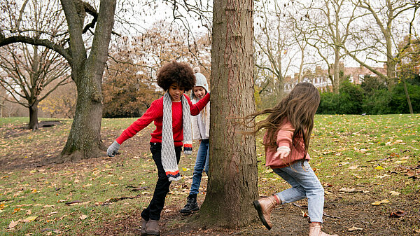 Three children playing outside. They are running around a tree in an autumnal scene.