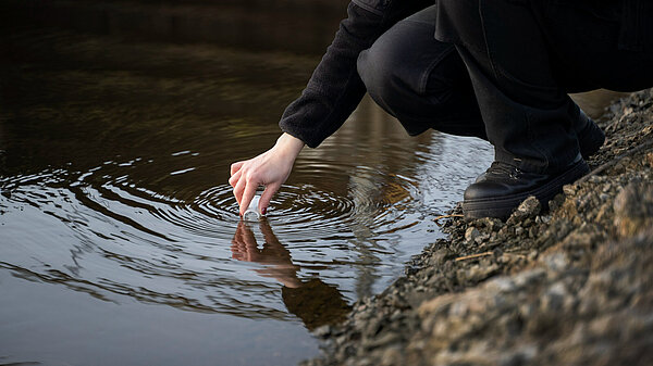 A human hand is holding a sampling bottle, taking a sample from a watercourse.