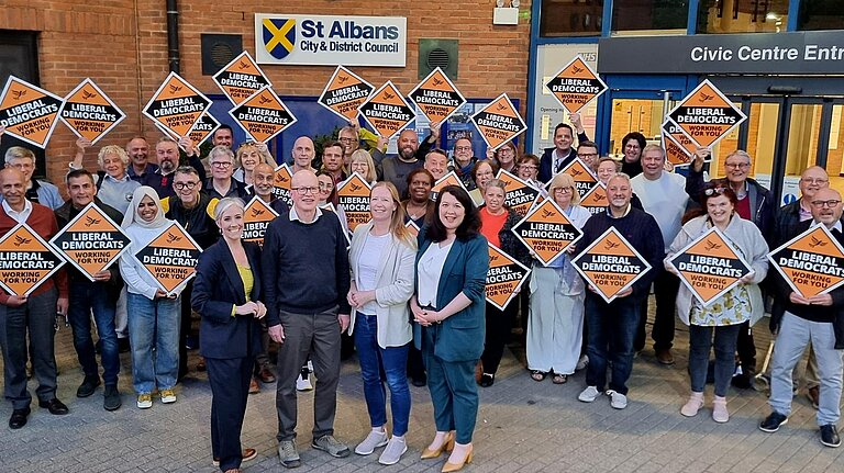 A group of Lib Dem councillors are standing in front of the St Albans District Council building, holding diamond shaped Liberal Democrat placards. Daisy Cooper MP, Councillor Paul de Kort, Councillor Helen Campbell and Victoria Collins MP (from left to right) are standing in front of the group.