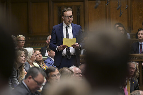 A man in a suit stands speaking in the House of Commons chamber, holding a piece of paper. Other MPs sit and listen around him.