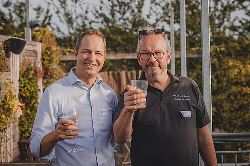 A photo of two men taken in a vineyard on a summer evening. On the left is Richard Foord and the right Ruud Jansen Venneboer. Both men are looking toward the camera and holding up glasses.