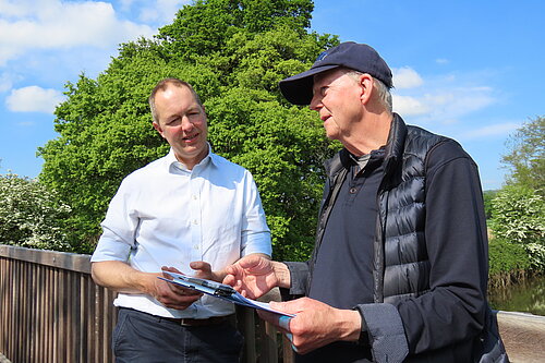 A photo of Richard Foord MP (left) speaking to a representative of the Otter Valley Association. Both men are stood on a wooden bridge with their backs to the railing. There is a tree behind them and the weather is clear and sunny.