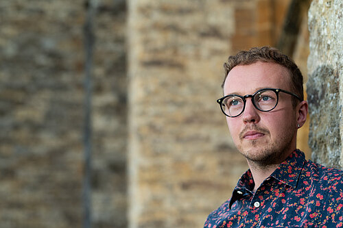 Adam Dance MP standing beside a stone wall, wearing glasses and a dark shirt with a red floral pattern, looking thoughtfully into the distance.