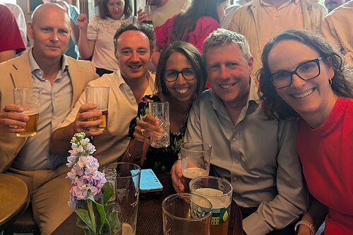Alison and other Liberal Democrat MPs in a pub watching the England Vs DRC football match, seated around a small wooden table, and other people behind.
