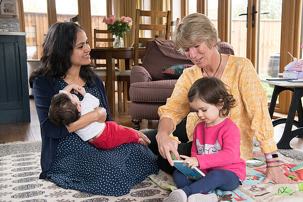 Pippa Heylings sat on the floor, looking at a book with a toddler, while the toddler's mother sits watching and holding her baby