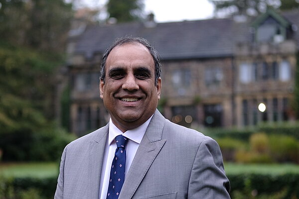 Cllr Shaffaq Mohammed. He is photographed outside in a garden area with a large stone house behind him. Shaffaq is wearing a grey suit with a blue tie, he is smiling and looking at the camera.