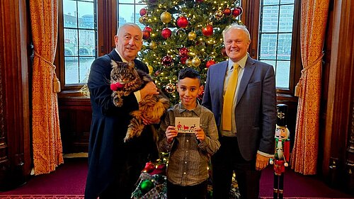 Clive Jones MP stood with local primary school student, Leo Sindhu, and the Speaker of the House of Commons, Sir Linsay Hoyle, in front of a decorated Christmas Tree. Leo Sindhu is holding his Christmas card design, which won the competition, and the Speaker is holding his large cat, named Attlee.