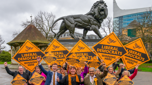 The Reading Liberal Democrats, cheering and holding orange diamond placards, in front of the Maiwand Lion