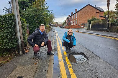 Cllrs Will Frass and Jane Brophy crouching next to a pothole on Deansgate Lanes