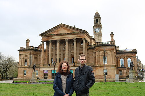 Anne Hannigan and Jack Clark standing next to each other, looking unhappy with Paisley Town Hall in the background.