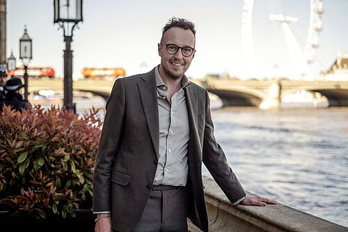 Adam Dance MP standing beside the River Thames outside Parliament, wearing a grey suit and smiling, with Westminster Bridge and the London Eye visible in the background.