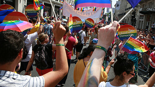 A Liberal Democrat group at Pride. The people are walking away from the camera, they are holding Lib Dem rainbow signs.