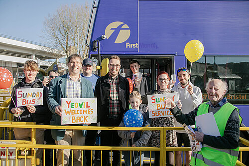 Group of people, including Adam Dance MP, smiling and holding colourful signs and balloons in front of a purple First Mendip bus, celebrating the return of Yeovil’s Sunday bus service.